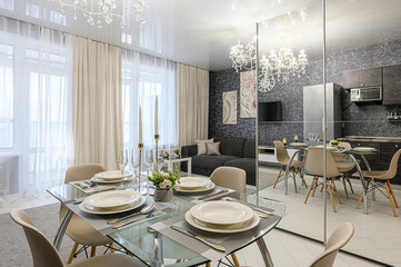A stylish dining area is set with a glass table, elegant plates, and tall candlesticks. A modern gray couch, a crystal chandelier, and neutral curtains add sophistication. A mirrored wall reflects