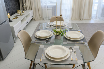 A neatly set dining table with plates, silverware, and placemats. The table is glass, and there are chairs around it. A small flower arrangement adds a touch of elegance