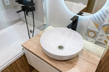 Contemporary bathroom with white bathtub, black shower fixtures on glass enclosure, round white vessel sink on oak vanity, LED-lit round mirror, abstract geometric tiles in gray, white, gold