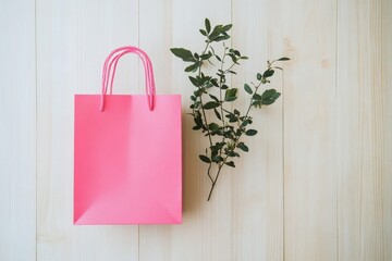 A vibrant pink shopping bag rests atop a light wooden surface, accented by a sprig of verdant foliage.