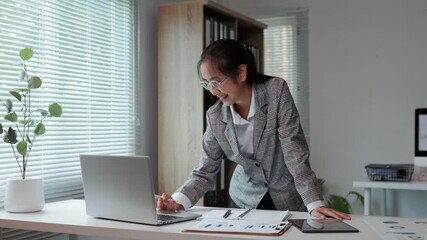 Professional businesswoman reviewing financial data on laptop and tablet, working strategically in contemporary corporate workspace with confident posture - Powered by Adobe