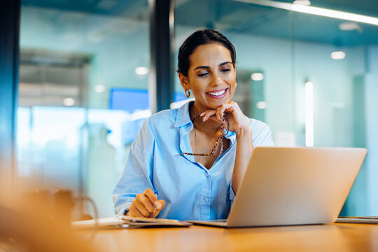 Fototapeta Smiling woman in office using laptop while holding glasses and working thoughtfully