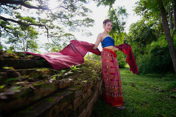 Woman traditional Thai attire stands gracefully lush garden, with sunlight filtering through trees. Her vibrant clothing and serene expression