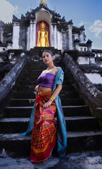 Traditional attire woman stands on ancient temple steps, golden Buddha statue in background. Cultural heritage and serene atmosphere