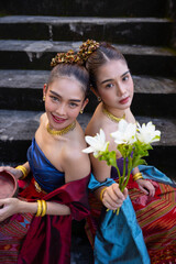 Two women in traditional Thai attire sit on stone steps, holding flowers and bowl, showcasing cultural elegance