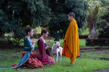 Two women in traditional attire kneel respectfully before young monk and dog in serene outdoor setting