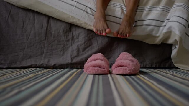Medium lockdown floor level shot of legs of unrecognizable young black woman with bright pedicure getting off bed, putting on pink furry slippers on stripy rug, then walking off