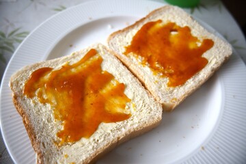Close-up view of two toasted bread slices with margarine and apricot jam on white plate.