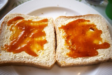 Close-up view of two toasted bread slices with margarine and apricot jam on white plate.