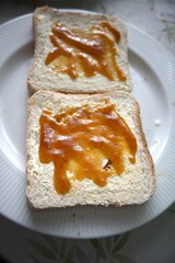 Close-up view of two toasted bread slices with margarine and apricot jam on white plate.