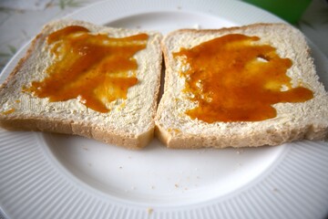 Close-up view of two toasted bread slices with margarine and apricot jam on white plate.