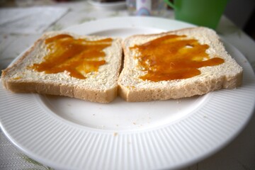 Close-up view of two toasted bread slices with margarine and apricot jam on white plate.