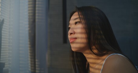 Young Woman Wakes Up Morning in Sunny Bedroom. Girl Sits Up in Bed With Sleepy Smile and Looks Out...