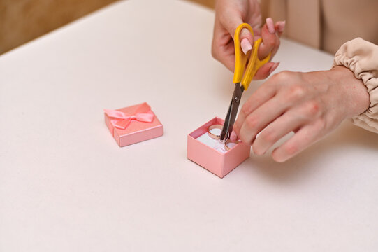 Hands cutting pink gift box ribbon for wedding rings, capturing engagement moment with delicate preparation. Scissors cutting satin ribbon of pink wedding ring box.