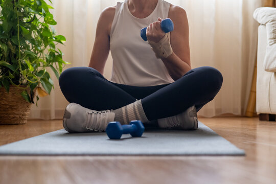 Mature woman lifting hand weights in living room, wearing wrist brace for safe home training and injury prevention during daily workout session