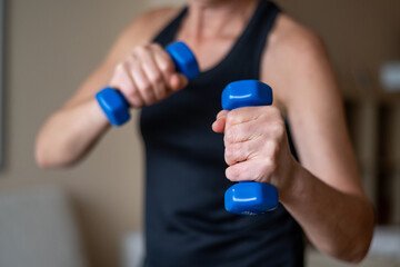 Faceless midlife woman performing resistance training with blue weights at home, routine fitness for health and balance