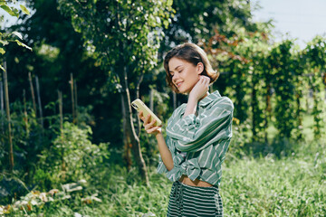 A woman stands outdoors in a lush garden, smiling softly while looking at her phone.
