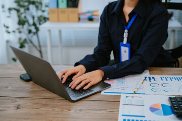 Young Asian businesswoman holding a pen and clipboard, analyzing statistical data on a tablet screen while working on a computer in a modern office environment