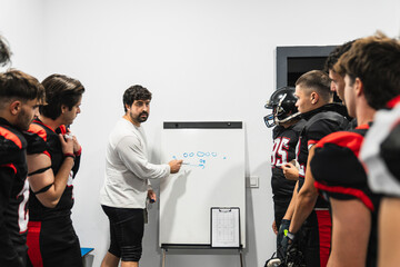 American football coach discussing game strategy on a whiteboard with his team in the locker room,...