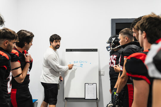 American football coach discussing game strategy on a whiteboard with his team in the locker room, planning for success