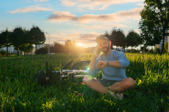A man sits cross-legged in a grassy park near his bicycle. He is talking on his mobile phone and looking at his smartwatch. The sun sets in the background