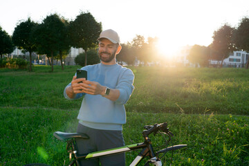 A smiling man in casual clothes and a cap holds his smartphone and takes a selfie next to his bicycle in a grassy park during golden hour. The sun shines brightly in the background
