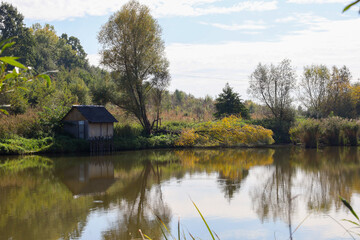 Obraz premium This autumn scene captures a tranquil forest pond reflecting the sky and the surrounding greenery. A cozy wooden hut stands near the water's edge under the shade of tall trees, creating a peaceful lan