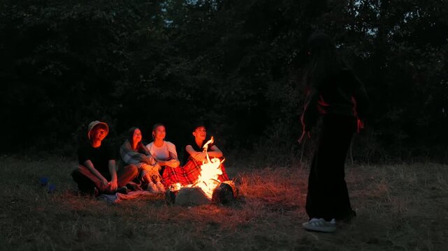 Teen girl telling stories to friends around campfire in the forest dusk
