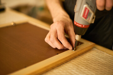 Man using electric screwdriver to fasten metal bracket on wooden frame, close up of hands performing assembly task on workbench, focus on manual craftsmanship