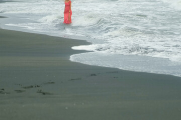 Woman in red dress enjoying sea vacations walking on black sandy beach in waves. Waves rolling...