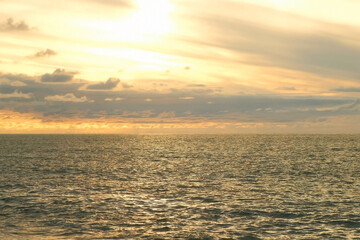 Golden sunset cloudy sky reflecting on calm ocean waters with ripples. Golden hour sunlight bathing calm ocean waves, soft clouds diffusing warm orange glow across expansive seascape. Calm atmosphere.