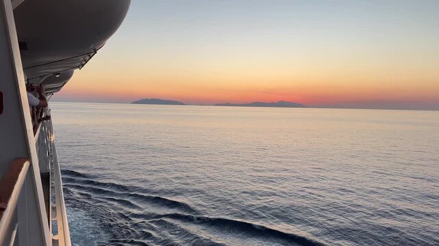 Forward tracking boat POV from a cruise ship deck sailing towards a colorful sunset over a calm sea and the islands of Sicily, Italy.