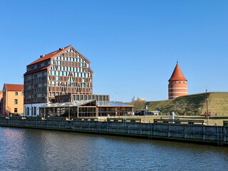 Panoramic view of the historical center of Klaipeda, Lithuania, featuring a traditional-style building with exposed wooden beams and a gabled roof. Red-brick castle's tower with a conical roof. 