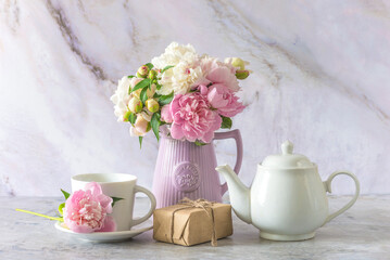 White tea pot, cup of tea, craft gift box and bouquet of pink and white peony flowers on a marbre background