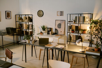 Modern office workspace featuring empty desks with documents, potted plants, shelving units with...