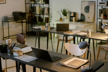 Modern office workspace showing empty desks with open laptops, documents, desk lamps and stationery...