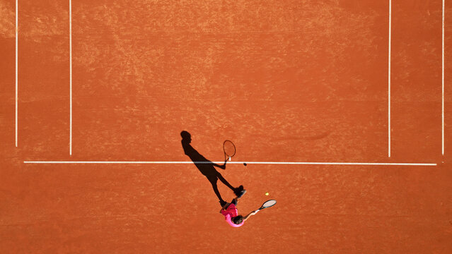 Drone shot of a tennis player in a pink shirt practicing on a clay court, mid-action with a dynamic shadow. Highlights sports training, skill development, and the textured orange surface.