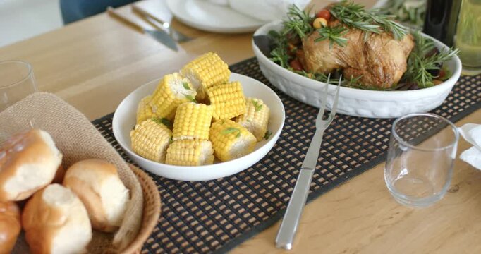 Camera is starting panning across dining table to reveal basket with rolls, corn, roast chicken