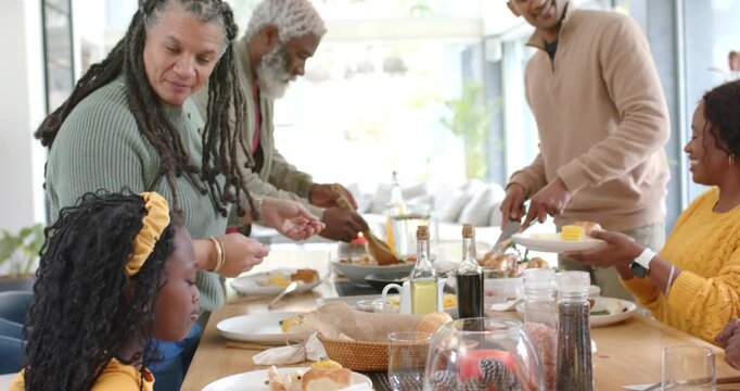 Diverse family passing dishes at wooden table when mature woman breaking bread for shared breakfast
