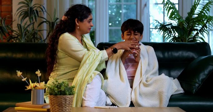Indian mother checking fever of son while sitting in modern home on sofa, taking care of his health, showing love, affection, parent child bonding, family lifestyle indoors, nurturing daily routine