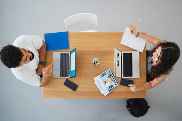 Two young adults, a man and a woman, are focused on their laptops and notebooks at a wooden table. They are engaged in a productive study or work session, demonstrating teamwork.