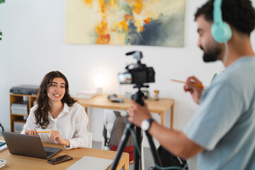 A young woman records a video at her desk, smiling and holding a pencil. A man with headphones operates the camera, filming her for an online content session.