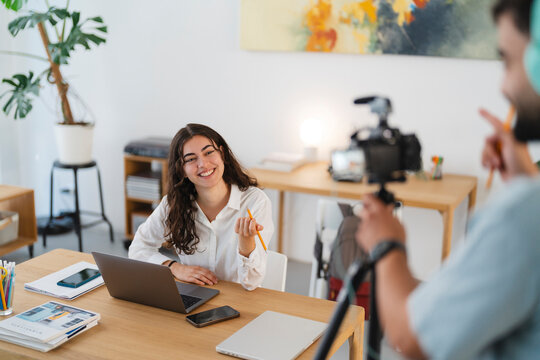 A smiling young woman sits at a desk with a laptop, holding a pencil, while a man films her with a camera. She is recording content for an online audience.