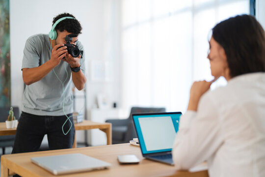 A male content creator with headphones films a female professional working on her laptop at a desk. They are engaged in a collaborative video production or interview in a bright office.