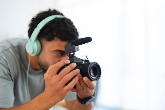 A young man with headphones records video using a camera with an external microphone. He focuses intently, capturing content for his project.