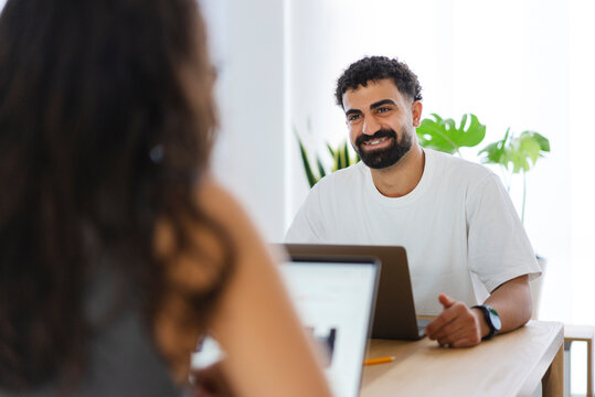 A smiling man with a beard and braces sits at a table with a laptop, looking at a colleague. They are collaborating or having a discussion in a bright office setting.