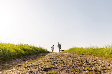 Fototapeta premium Under a clear blue sky, a man and his son walk together with their dog along a gravel path surrounded by tall grass. The scene captures a moment of family bonding outdoors