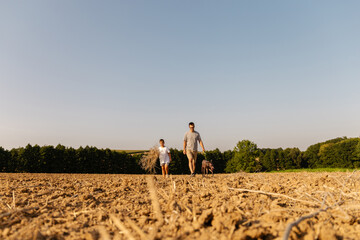A man and his son are walking together in a dry field, accompanied by their dog. The sky is clear with a warm sun shining down, creating a peaceful atmosphere
