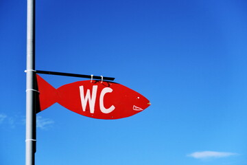 WC. Panneau rouge peint en forme de poisson sur fond de ciel bleu.