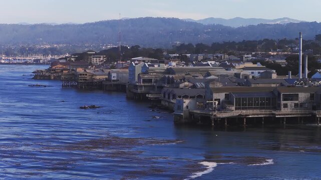 Aerial view of Monterey, California, showing Cannery Row, waterfront buildings, kelp beds, a marina with boats, and forested hills in the background.